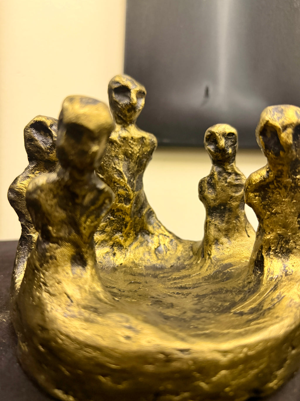 A close-up of a textured gold sculpture depicting five stylized, elongated human figures standing in a shallow basin, with a blurred indoor background.