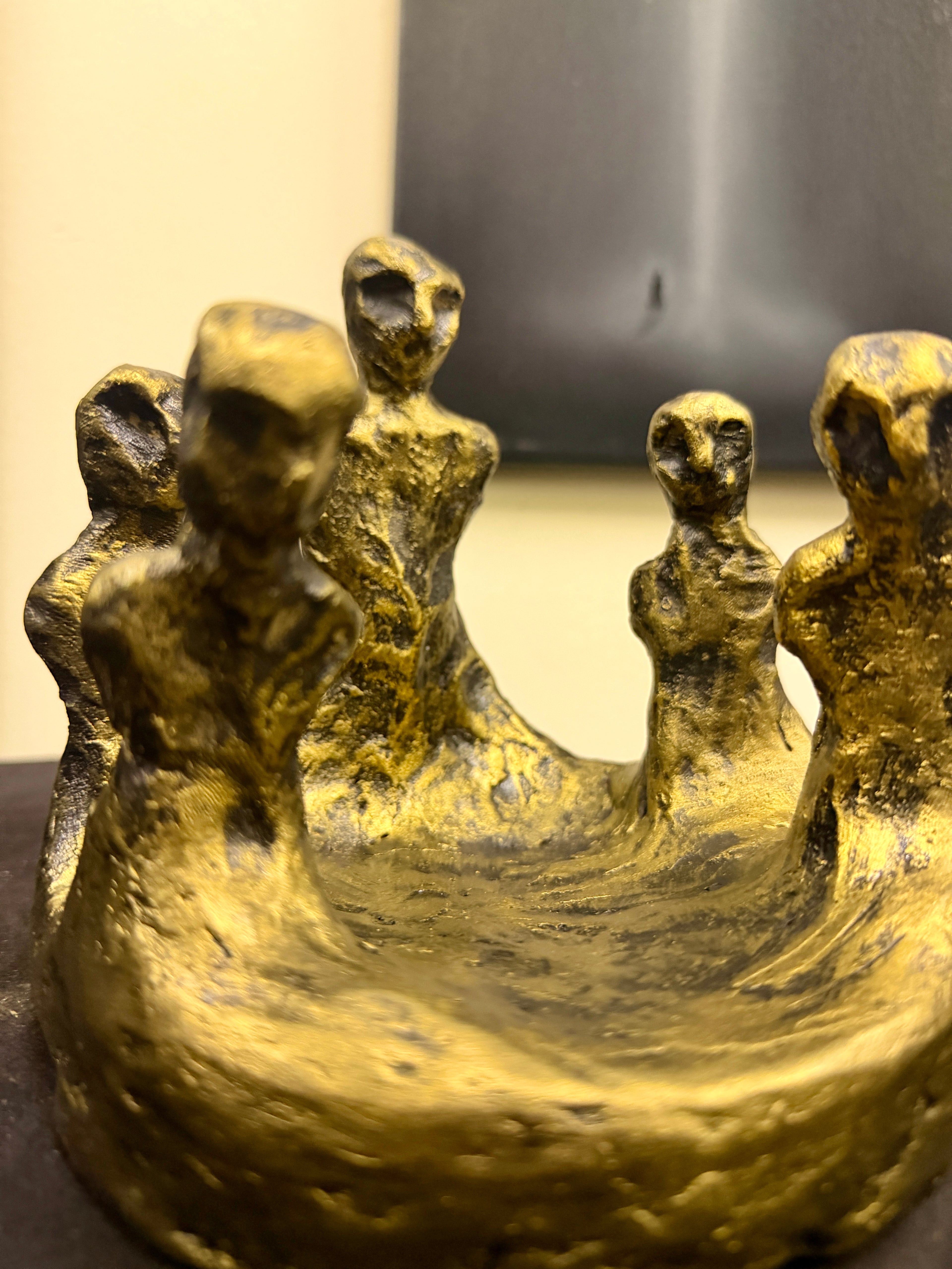 A close-up of a textured gold sculpture depicting five stylized, elongated human figures standing in a shallow basin, with a blurred indoor background.