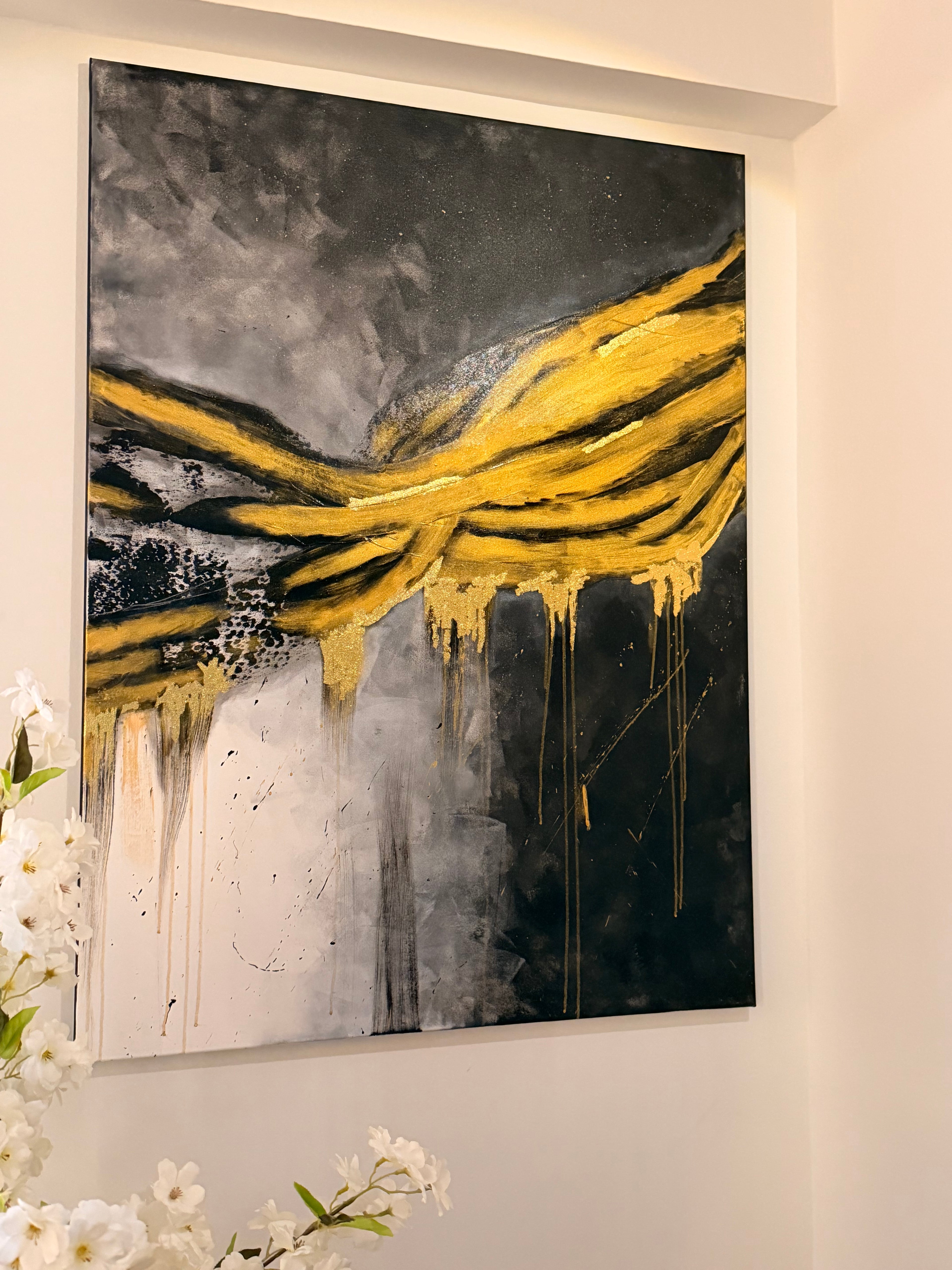 An abstract canvas painting featuring bold yellow paint drips and layered brushstrokes over a dark gray, black, and white background, displayed on a white wall with white flowers in the foreground.