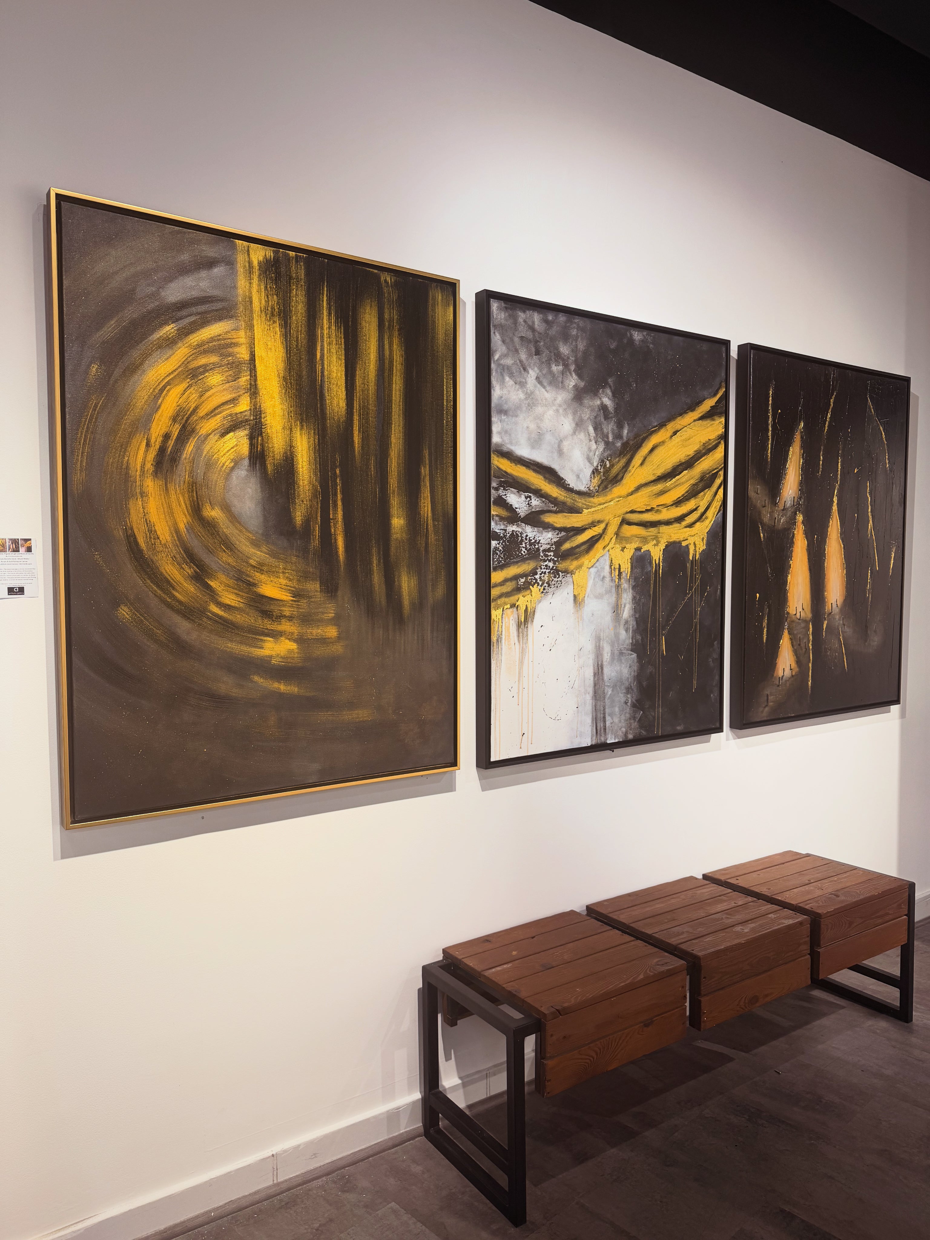 Three framed abstract paintings with bold yellow and black brushstrokes displayed on a white gallery wall above a wooden bench.