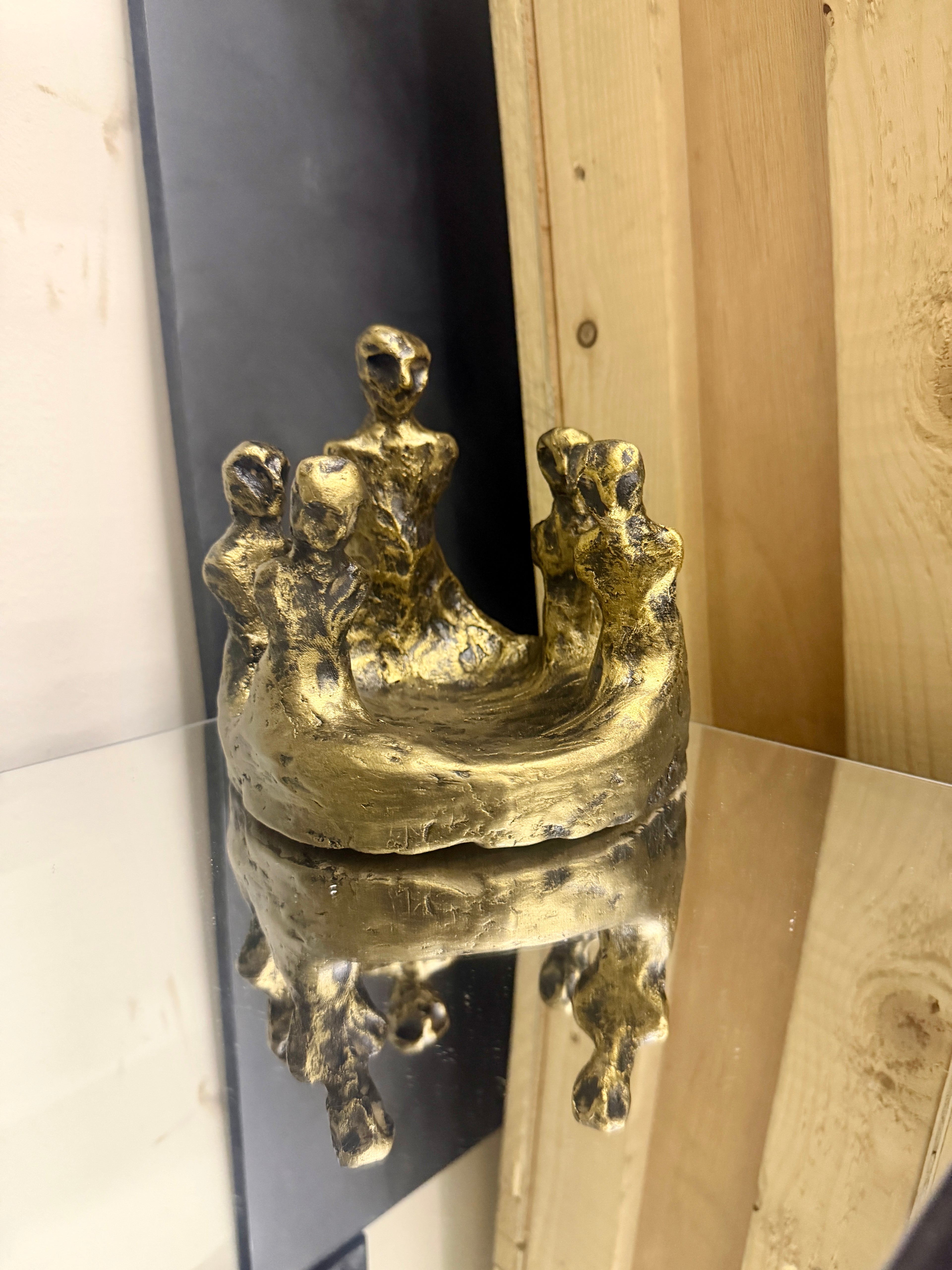 A small abstract metallic sculpture in a gold finish, displayed on a reflective surface with a wooden panel background.
