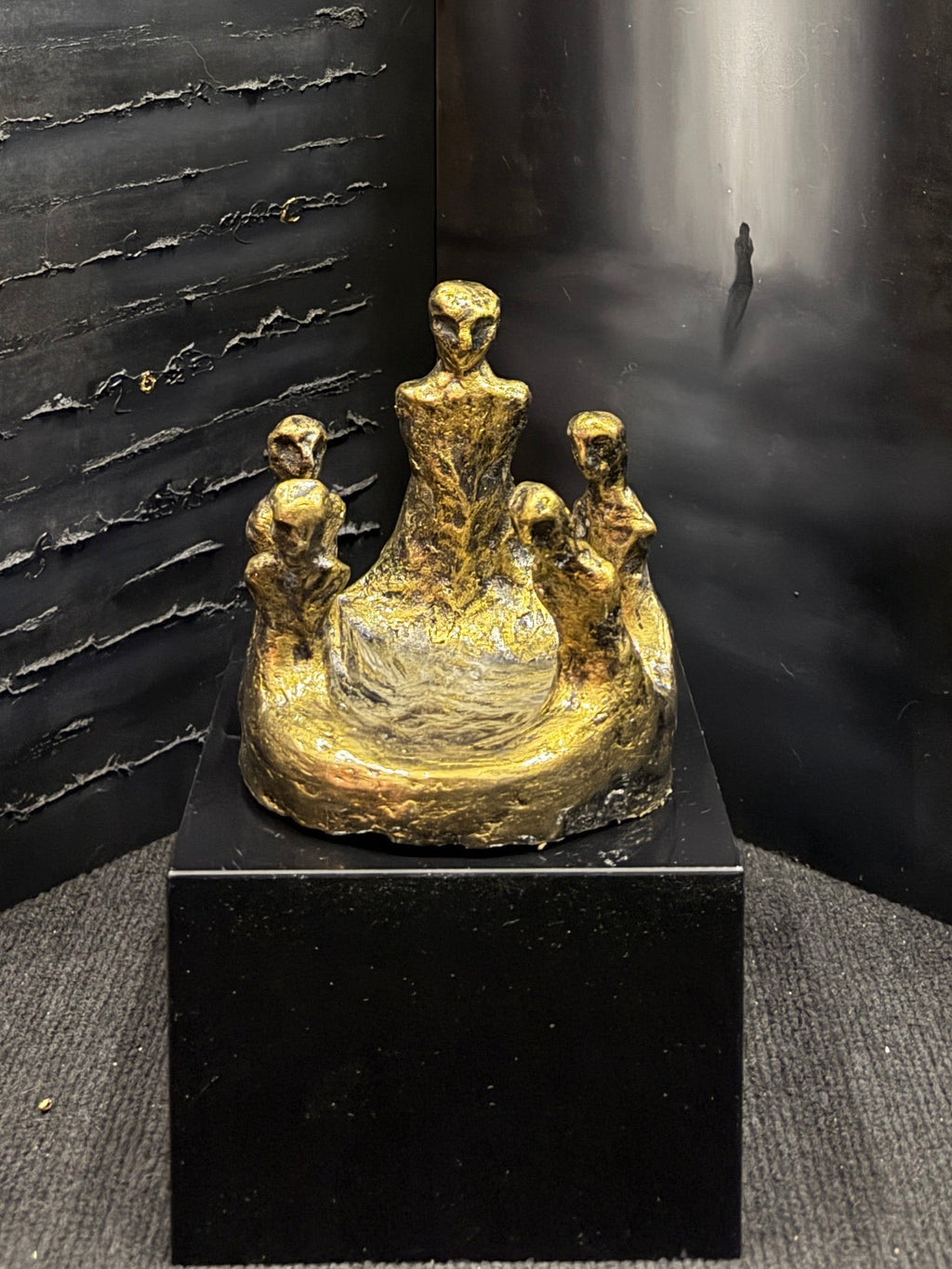 A dark, moody art display featuring a large three-panel folding screen with textured black and gray panels, one panel showing a misty white vertical light. In front of the screen sits a small gold-toned figurative sculpture on a black pedestal.