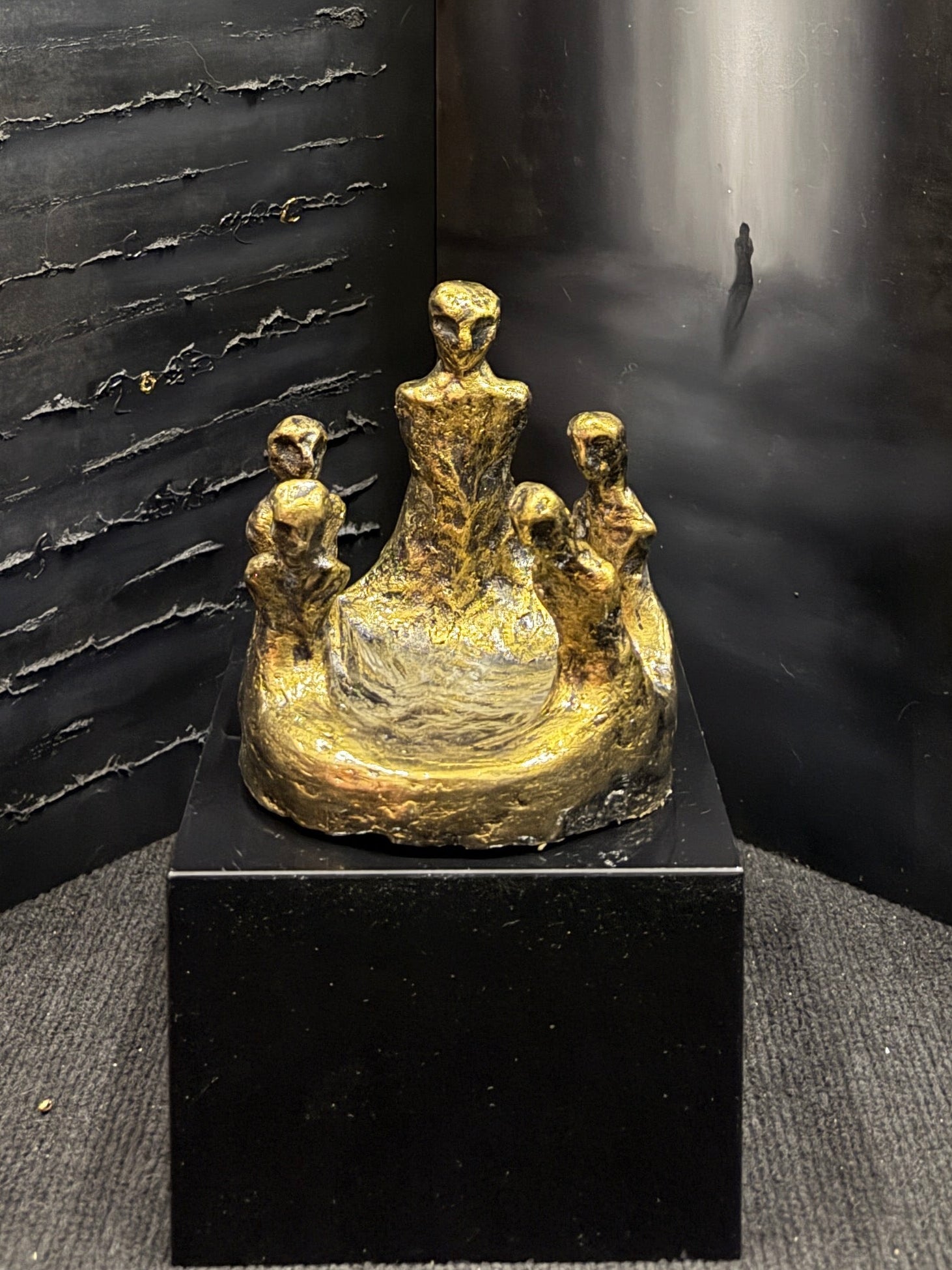 A dark, moody art display featuring a large three-panel folding screen with textured black and gray panels, one panel showing a misty white vertical light. In front of the screen sits a small gold-toned figurative sculpture on a black pedestal.